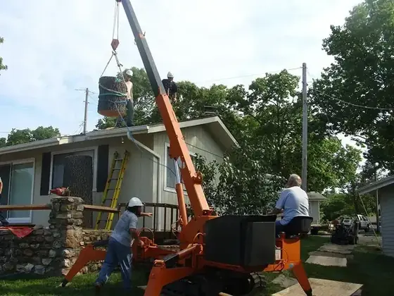 An orange spider lift and crane lifting a cut trunk section clear of a lake house roof